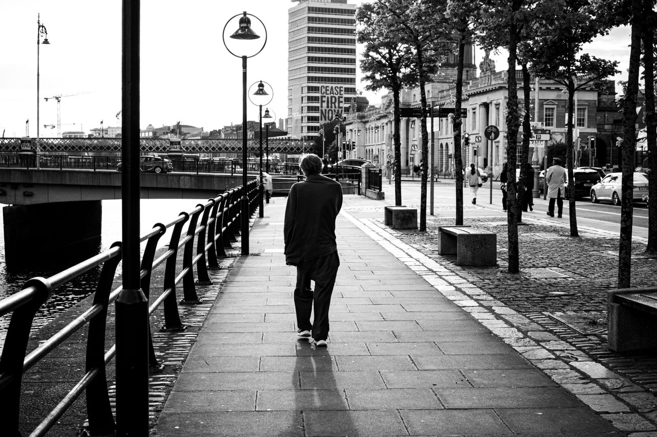 Black and white photo of a solitary figure walking along a riverside street in Dublin, Ireland.