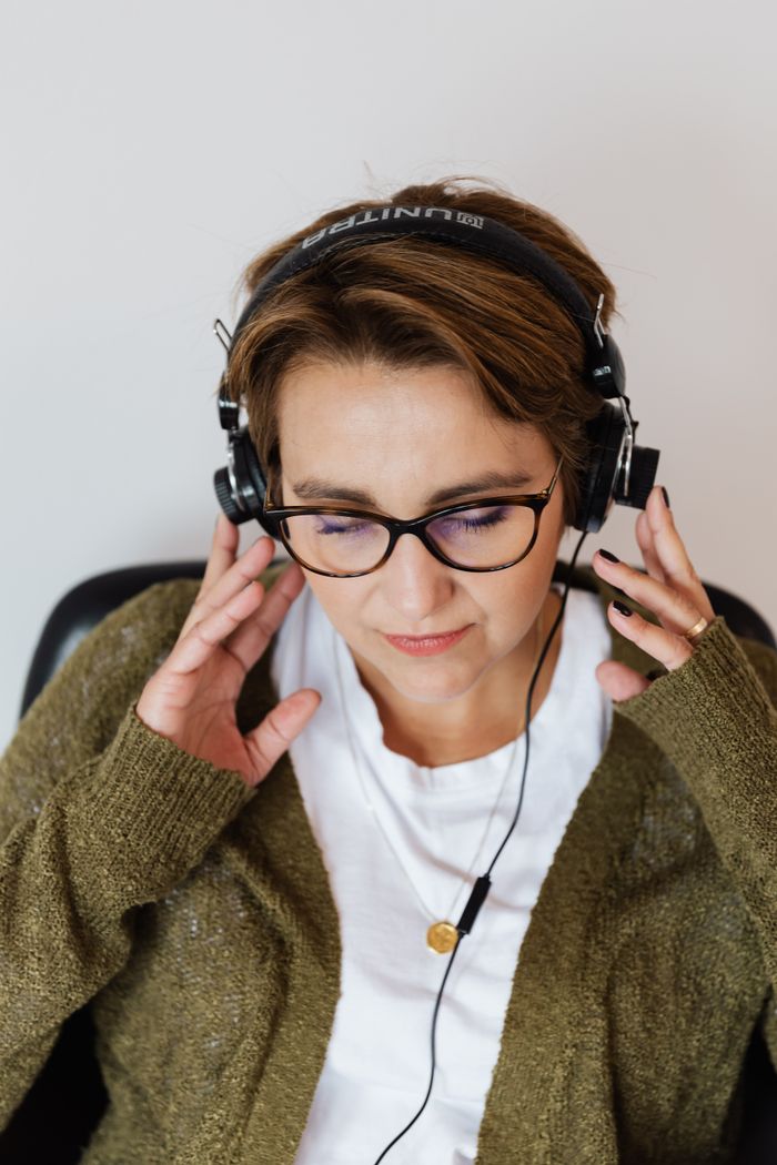 Home Glad relaxed female wearing eyeglasses and warm casual clothes resting on chair with eyes closed and listening to good music via wired headphones on white background